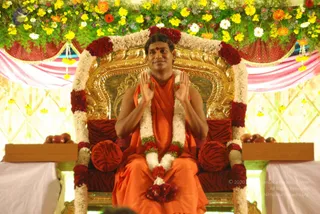 A man in an orange saffron robe sits on an ornate gold throne upholstered in red with a striped pattern, decorated with garlands of white and red flowers. His palms are pressed together in a namaste gesture and he smiles. Above him hang an overhead canopy of yellow, orange and white flowers and pink and red draped cloth with gold tassels. Flanking him on small wooden stands are plates of red fruit, and a white cloth covers the stands. The scene appears to be a darshan setting.