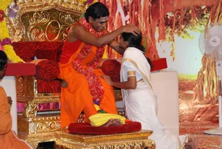 A man in saffron-orange robes with a red and pink flower garland sits on an ornate gold-and-red throne, placing his hand on the head of a woman kneeling before him in a white sari with gold border. A second person in orange robes sits at the lower left edge. Behind them is a large backdrop of a sacred tree trunk and a fan stands at right.
