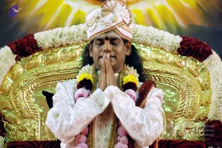 A seated man with eyes closed and palms pressed together in namaste sits on an ornate gold throne with carved scrollwork. He wears a white and gold turban with a fan-shaped ornament, a red bindi, and a white embroidered silk kurta with a dark ornamental sash. Garlands of white jasmine, red roses and yellow marigolds drape the throne and his neck, and a glowing yellow-orange backdrop fills the background. The setting suggests a formal seated darshan on a throne.