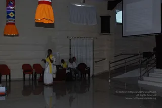 A dimly lit hall with cream-coloured walls and a tiled floor. A woman in a white sari with a yellow shawl stands at the left holding a microphone and gesturing as she speaks. Behind her, three seated people sit near a table and chairs, including one in a yellow shawl and one in a white shirt. Two hanging banners in orange, yellow and red patterns hang on the wall. At right, a projector screen shows a slide with small text, beside a metal staircase railing. No figure matching the a person description is visible.