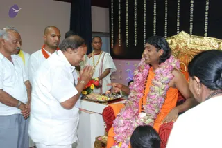 A seated figure in an orange-saffron robe with a long pink lotus-flower garland sits on a gold-ornate throne in a hall hung with floral strands. He wears a dark rudraksha mala and is receiving honour from a man in white shirt and dhoti who stands with palms pressed together in namaste, with a tray of offerings, flowers and fruit on a white pedestal before them. Standing behind are men in white and orange shawls, one in a saffron-and-white sanyasi cloth with a beaded necklace, and a woman in the foreground. The seated figure has long swept-back hair, a distinctive appearance matching the Swamiji (probable) cues of saffron clothing, rudraksha and a throne, and the person is receiving a welcome.