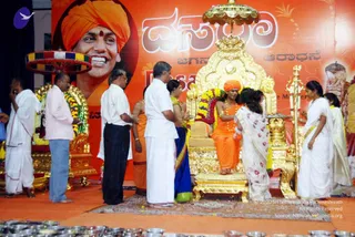 A photograph of a Dasara celebration on a stage with an orange-red backdrop showing a large portrait of a smiling man in an orange turban with a red bindi, and Kannada and English lettering reading Dasara. A seated figure on an ornate gold throne with a gold canopy wears an orange-saffron dhoti and cloth, an orange turban, and an orange rudraksha-style bead necklace, with flower garlands draped over the throne; a woman in a white sari with a red-patterned border stands at the right holding a tray. Around the throne, several people in white dhotis and shirts, a woman in a blue sari, and a woman in white stand attending. Brass plates or bowls with offerings sit in the foreground, and a gold-handled staff is held beside the throne.