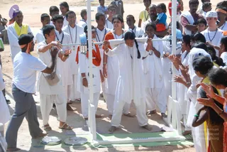 Outdoor photo of a youth event on a dusty ground with a crowd of young people in white kurtas and white dhotis or salwar-style trousers, many wearing blue lanyards and some orange sashes. In the centre, a young woman in white with a long braid and white shawl bends forward under a horizontal white metal bar balanced across her shoulders on a white stand, while others around her clap and cheer. A man in a white t-shirt and grey trousers with an orange headband stands at left gesturing. Spectators in the background watch, and a child is held up at upper right.