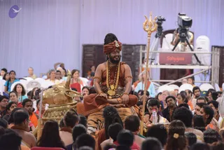 A man with long dark hair and a full beard sits cross-legged on a tiger-skin-draped seat, smiling, in a saffron-orange turban and wrap with a brown-and-orange shawl, wearing a heavy gold necklace, rudraksha-style beads and gold armlets. He holds a tall gold trident beside him, and a golden bull figure stands at his left. A large crowd of devotees sits in front of him, many in white and orange, while camera operators on a scaffold stand behind. A banner reading Sadyogata hangs on the platform. a person appears in person, seated on the throne-like seat in a satsang setting.