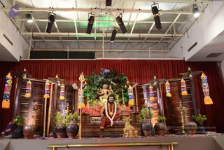 A photograph of a stage set for a satsang or darshan. A man with long dark hair and a full beard sits on an ornate carved wooden throne in orange-rust robes, with a white flower garland and a rudraksha-style mala around his neck, his arm resting on the armrest. Behind the throne stands a golden deity murti of a seated figure with a trident beside it and green potted foliage. Dark red curtains hang behind, with wooden panels flanked by decorative colourful fabric tassels. Brass oil lamps, terracotta pots with plants, a golden animal figure and a microphone stand surround the stage, and stage lights hang from the white ceiling.
