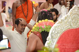 A photograph of a ritual in an ashram hall. In the foreground a man in a white shirt and white trousers stands with one arm raised, holding an orange fruit aloft, and his other hand pressed to his forehead with eyes closed. Beside him a man in an orange robe and a dark wooden bead mala stands. In the centre, a figure seated from behind with long dark dreadlocked hair wears a headdress of yellow citrus fruits, pink and red fruit slices and pink flowers, sitting on an ornate silver and red upholstered throne. A man with a beard and other white-clad devotees are in the background, along with a silver stand.