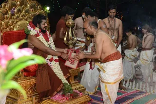 A seated bearded man in saffron-orange robes sits on a golden ornate throne with a red cushion, wearing a fresh garland of white flowers with red roses, while a bare-chested priest in white dhoti with an orange-red waist cloth and rudraksha mala bends forward to offer him a red-and-white bundle. Behind them a gold trident stands near a brass vessel, and other shirtless men in white dhotis, some with sacred ash on their foreheads, stand or sit in the background with flower petals on the ground. Strings of lights glow in the dark night setting.