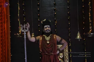 A man stands in front of a dark backdrop strung with small lights and hanging brass bells, smiling and raising his right hand while holding a silver trident. He wears a crescent-moon crown over his dark hair, white forehead markings, a heavy gold necklace, layered rudraksha malas, and a deep maroon-red garment with gold border. Another gold-framed decorative panel stands to his right.