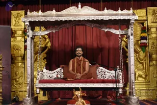 A seated man in saffron-orange robes sits on a silver-framed swing-style throne with red cushions and carved silver railings and chains hanging from the canopy. He wears a red bindi, a saffron shawl draped over his shoulders, and a gold-toned necklace, with a microphone stand positioned beside him. Behind him hang deep red curtains flanked by ornate gold-painted carved panels showing figures and animal motifs. A small gold figure of a bull, a nandi-style statue, rests on a red cushion in the foreground, and a multicoloured tassel hangs at the right edge. Oil lamps stand at the left edge. The setting is a satsang-style stage with a ceremonial seat. Swamiji (probable) appears in saffron robes with a rudraksha-style necklace and a long swept-back hair, seated on the throne.
