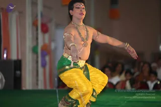 A photograph of a performer on a green stage mid-dance, bare-chested and wearing a yellow and green dhoti with gold waistband, multiple beaded necklaces, gold armbands and bangles, and a tilak on the forehead with painted eyes and red lips. His arms are extended outward and one knee is lifted. Behind him are hanging orange, green, purple and red decorations and a blurred audience of seated people in white and colourful clothing.