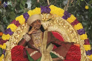 A seated man sits on an ornate gilded throne with a red cushion, backed by a garland of yellow and purple flowers and leafy branches with small orange fruit. He wears a cream turban, a red and gold brocade kurta with embroidered cuffs, heavy gold necklaces, a green and cream shawl draped over his lap, and a red bindi on his forehead. He rests one hand against his cheek and smiles. a person: the seated figure's long swept-back hair, turban and ornate gold and brocade attire match the cues, and the page indicates his presence, so the appearance-based label applies.