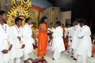 A photograph inside a hall with a golden throne decorated with yellow, white and red flower garlands behind a central stage. A long-haired man in a flowing saffron-orange robe with a gold-trimmed border stands centre-right, smiling, extending a white gift bag to a man in white ritual clothes and a rudraksha mala. Several men in white dhotis and shirts, some wearing red-brown rudraksha malas, stand in a row holding white cloth bags with tree prints, and an assistant in orange clothing and other people watch from behind. The stage has a red floor mat and maroon and orange drapes.