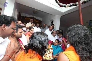 A courtyard scene in a white-walled house with a red-and-white tasselled canopy overhead and a floral garland hanging on a doorway. In the foreground, two women in orange saris with long dark hair face away, one holding a silver plate of flowers and fruit. A bare-chested man with a rudraksha mala and a red thread on his wrist stands at left beside a man in glasses. A woman in a teal sari holds a pink garland, while a crowd of men in white shirts and dhotis stands behind. No one visible clearly matches a distinctive appearance cue, so no identification is made.