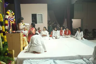 Indoor ceremony on a raised white-covered platform. On the left, a figure with long swept-back dark hair, wearing white robes and a white garland, sits on an ornate golden throne-like seat beside a floral arch of marigold and white flowers and a small offering tray of fruit and flowers. Facing this seat, a woman in an orange-red sari with a bindi stands with palms pressed together in namaste, and a figure in white sits with head bowed on the platform. Seated devotees in white and orange-red shawls line the platform, and more people stand in the background near dark drapes.