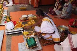 A priest in white dhoti and shawl with gold border sits cross-legged on an orange mat, pouring from a small brass vessel over a ritual setup of silver trays, brass bowls of yellow flowers, turmeric-coloured sweets, a coconut and mango leaves, and a folded green cloth. Two seated couples in the background wear flower garlands: a woman in a maroon sari and a man in a grey shirt. The scene suggests a puja or abhishekam arrangement. No clear a person candidate is visible.