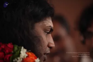 Close side profile photograph of a man with long dark hair swept back, a light vertical marking on his forehead, and a garland of red roses, orange marigolds and white flowers draped over his shoulder. His eyes are lowered and his expression is calm. The background is blurred warm brown with two out-of-focus figures behind him.