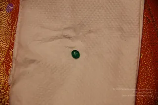 Close-up photograph of a single small, smooth oval green gemstone, a cabochon, resting on a white textured cloth with a woven pattern. The cloth is draped over a red fabric with gold metallic brocade patterning at the edges. No people are visible.