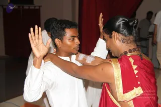 A photograph of two people facing each other in close physical contact during what appears to be a hands-on practice session inside a hall with a deep red curtain behind them. A young man in a plain white kurta has his right palm raised, while his left arm rests on the shoulder of a woman in a red sari with a gold border, her hair in a bun and a rudraksha mala around her neck; she raises her own hand toward his. A bandage-like white patch is visible on the man's upper arm. Another person in white stands partly visible behind them, and several people in light clothing are at the right edge. No deity, fire or ritual objects are visible.