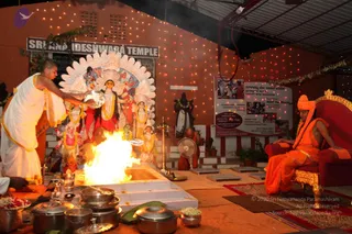 Night-time photograph at Sri Nideshwara Temple. A priest in white dhoti and shawl with a bare shaved head stands at left holding a brass vessel over a bright fire pit (homa or yagna) in front of a deity murti decorated with white floral garlands and an ornate white-and-red backdrop. Another orange-robed figure sits behind the fire, and metal offering pots, bowls, and a plate of flowers rest in the foreground. At right, a figure in orange robes with an orange turban, rudraksha mala and a shawl sits on a red-and-gold ornate throne on a rug. Strings of lights decorate the orange walls and a corrugated roof above. A signboard reads Sri Nideshwara Temple, and a panel nearby bears photos and text about Meet the Master and admission.