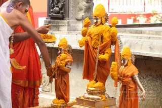 A photograph of a ritual turmeric anointing of three metal deity murti standing on a silver-toned base before a dark stone wall. The large central murti wears an orange garment and a tall yellow crown, its arms and body coated in yellow turmeric paste. Two smaller murti flank it, also draped in orange cloth and covered in yellow turmeric. On the left, a shaved-headed priest wearing a white dhoti, an orange-red shawl and gold armlets and necklaces holds a metal plate of yellow turmeric and applies it with his fingers. Yellow turmeric is visible on his hand and the murti.