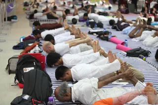 A photograph of a yoga session in a large hall, with many participants lying prone on striped blue-and-white mats in forward-bending poses, arms stretched forward and faces down. Most wear white clothing, with a few in darker or orange garments. Backpacks, water bottles and pink yoga bolsters lie beside the mats along the marble floor, and queue barriers line the left edge. The participants are many and only partially visible, so the count is best given as a range. No individual is clearly identifiable as a person.