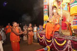 Night photograph of a rathotsavam procession. A seated figure in orange silk with a gold crown, heavy gold necklaces and a sword held upright rides an ornate golden chariot decorated with colourful patterned panels, red and yellow garlands, and a blue-clad figure on a platform above. In the foreground a priest in orange robes and beads holds a lit aarti lamp with flame, while a second priest in orange stands beside him. Devotees in white and orange stand with hands pressed together along the road, and smoke drifts through the dark background.