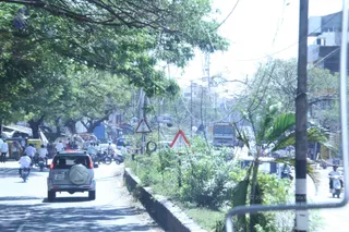 A busy tree-lined street seen from inside a vehicle, with a silver SUV with a spare tyre mounted on its rear driving ahead. Several motorcycle riders wear light shirts and helmets, and pedestrians walk along the left edge. Red-bordered triangular road warning signs stand near the roadside, and a large bus is visible further up the road. Overhead power lines cross the scene, and green shrubs and palm plants line a low concrete kerb on the right. Buildings and a tall pole fill the background under hazy daylight.