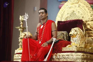 A man seated on an ornate golden throne carved with lions and a large arched back, wearing a red saffron-toned wrap over the shoulder, red dhoti-style lower garment, multiple rudraksha and bead malas, a blue beaded necklace and a ceremonial white pendant. He has a white tripundra forehead marking with a red dot, a small lapel microphone, and holds a silver trident in one hand and a white-wrapped staff in the other. Behind him are a gold oil lamp and red draped curtains. Swamiji (probable) appears here given the presentation cues: long swept-back hair, ornate gold-and-bead adornment, saffron-red clothing and rudraksha malas, seated on a throne as a teacher giving a discourse.