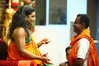 A photograph of a seated person with long, swept-back dark hair wearing an orange-yellow saffron-toned sari with green blouse and a rudraksha mala. The person hands a small white card or paper to a smiling man in an orange saffron shawl over a white shirt. Behind them is a floral garland and gold ornamental decoration, and a partially visible person sits at the lower right. The setting appears to be an indoor satsang-style room.