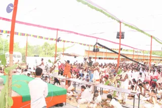 Outdoor pandal ceremony under a white canopy with pink, green and multicoloured bunting and orange-painted poles. Several priests in white dhotis and bare-chested or white upper cloths perform a ritual at a long table, one holding a brass aarti lamp, another with a kalasha-like vessel. A man in an orange robe stands centre-left, and a bald man in white stands at the table near a video camera on a boom. Large crowd of seated devotees in white clothes fills the foreground and rows, with plastic chairs at the right. A green-draped raised platform with banana leaves and fruit sits at left. Trees and a building are visible beyond the tent.