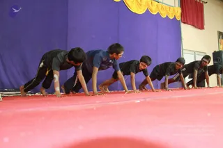 A photograph of a group of young boys in dark athletic clothes performing a synchronized plank or push-up exercise on a red stage floor. They are barefoot, with hands planted and bodies in a straight line, facing the same direction. Behind them hangs a purple curtain with a gold-yellow valance, and a red curtain and a framed picture appear at the upper right. The boys wear black or navy tracksuits and shirts, some with white marks on their arms.