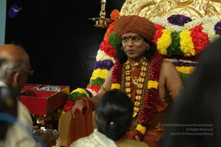 A seated man in a rust-red turban and orange-saffron robe sits on a throne-like seat decorated with garlands of red roses, yellow and white flowers, and green leaves, with a gold-framed backrest behind him. He wears a heavy gold necklace, a red-and-pink flower garland, gold armlets and earrings, and a red tilak on his forehead, and he is speaking while one hand rests on his robe. In the foreground, a bald older man in glasses and a person with dark hair in a bun, dressed in white, are partly visible. A red lacquered box holding small silver items and a brass oil lamp stand nearby, and a brass kalasha-style lamp rises at the top left. Probable Swamiji by his turban, saffron robe, gold necklaces and garland, seated in a position of honour.