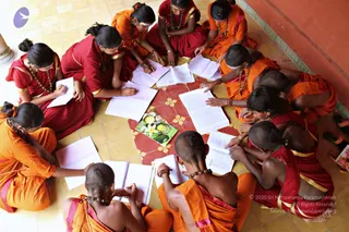 Overhead view of a circle of young students seated on a yellow tiled floor around a red-and-gold patterned tile, writing in notebooks and loose papers. Several wear saffron-orange robes with orange shoulder wraps and rudraksha malas, while others wear red and gold garments; most have shaved or closely cropped heads and some have hair pulled back. One student holds a pen and writes, and a glossy magazine with a lemon and fruit cover lies on the tile at the centre. Beside them is a pale wall and a column at the edge of the frame.