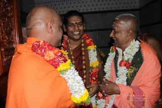 Three men stand close together in an indoor setting with a dark wall and an ornately carved wooden panel at left. The man at left, shaved-headed and seen from behind, wears an orange saffron robe and a garland of red and yellow flowers. The central man, smiling broadly, wears a maroon robe, a dark red-brown bead necklace and ornate gold-toned beads, a red bindi on his forehead and a garland of red and white flowers. The man at right, short grey hair, wears an orange robe over a dark green patterned shawl and a white garland of jasmine-like flowers with red blooms. They clasp hands and greet one another warmly.