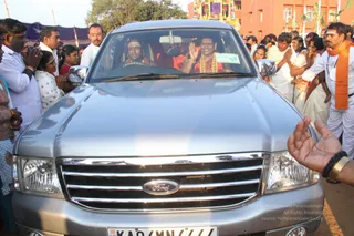 A silver Ford SUV drives through a crowd outside a temple compound with flags and decorative bunting. In the front seats, a driver in a white headband and a passenger in a red-and-orange garment wearing a gold necklace and a flower garland wave to the crowd. Many devotees in white and cream clothes, some with folded hands in namaskar, press close to the vehicle on both sides. A man in white with an orange shawl stands at the right edge, and a hand is raised in the foreground. Red brick buildings and a purple tent frame the background.