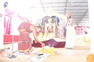 A photograph of a ritual at a pavilion with a red and white draped backdrop and corrugated roof. In the foreground, a man in a maroon robe with gold borders bends forward over a yellow-and-gold box on the floor, placing offerings and smoke rising from a small fire. Behind him, a figure in a white dhoti with a saffron head wrap and garlands stands with hands folded in prayer. Behind them, a gold-decorated seat with a red velvet base holds two garlanded deity murti figures with gold drapery and a blue canopy. Brass plates of flowers and small vessels sit on the floor. Two people appear in the background on the right. The image is washed out and overexposed.