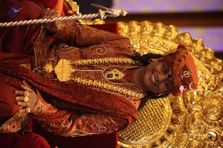 A smiling person in a rust-red embroidered tunic with gold zari work and jewelled beadwork sits on an ornate golden throne. The figure wears a matching red-orange turban with a gold crescent-moon ornament and a jade-green pendant on a gold necklace, and a red-orange shawl drapes the shoulders. A silver ceremonial staff with a trident-like top rests at the upper left, and a plain red seat cushion appears at the lower left. The image is rotated sideways. a person: the long swept-back hair, ornate gold-trimmed clothing and crown-like turban are consistent with the page's identification, but the appearance is not conclusive.