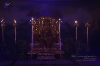 A dimly lit stage set at night-like blue lighting shows a seated figure on an ornate gilded throne with a large golden circular backdrop. The figure wears a saffron-orange robe and a beaded necklace, with a belt-like waist sash, and is seated among tall silver trishula staffs and dark foliage. A painted mountain and cloud backdrop with a trident motif fills the background. The seated figure appears to be a statue or effigy rather than a living person, so the scene reads as a darshan tableau for Kalabhairava. a person cues are present only in the seated figure's saffron robe and beaded necklace, and the figure is a depiction, not a physically present person.