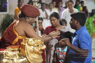 A photograph of a seated figure in a red-maroon turban, orange-red garment with gold border and a rudraksha mala, extending a hand in blessing toward a young girl in a striped teal top with a conference-style badge. A man in a blue polo shirt leans in beside her, and a woman in a red patterned blouse and white shawl watches. Behind them, several women in white and colourful saris stand near a carved golden lion-headed throne armrest, with a chrome stand and green plants in the background.