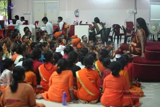 A photograph of a gurukul session in an indoor hall with pale pink walls and a fan. A long-haired figure in saffron-orange robes sits on a red cushioned seat at the right, holding a microphone and addressing a seated group. Many children and young people in orange and saffron clothing, some with white or orange head cloths and beaded necklaces, sit cross-legged on the floor in rows facing the speaker. Some are dressed in white and pink. Behind them, staff sit at desks with computers and paperwork, and a red chair and a water bottle are visible in the foreground.
