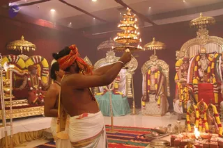 Indoor ritual scene in a hall with maroon walls and a warm-lit ceiling. In the foreground a bare-chested priest in white dhoti with an orange-and-red head cloth raises a tiered brass aarti lamp with many lit flames, while a second priest in white stands beside him. To the right stand deity murti and figures decorated with yellow and orange flower garlands, on an ornate silver-framed altar; a small fire pit with offerings and brass vessels sits on the floor at right. On the left, a seated figure in orange-and-red robes and an orange head cloth sits on a gold-framed throne draped with garlands, with a golden parasol above. Several empty decorated thrones with umbrellas stand across the hall. The scene shows a puja with aarti being offered.