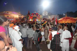 A night photograph of a crowded street procession with bright floodlight overhead. Several large ceremonial umbrellas dominate the frame: one in red, gold and blue with white dove emblems and Hindi/Sanskrit lettering, and a larger red-gold umbrella at right. Red and saffron flags rise above the crowd. Many people in white dhotis and kurtas, some wearing orange and white turbans or head cloths, walk alongside saffron-clad sadhus and a woman in a red sari. A man at lower right wears a white face cloth and a black jacket. Below, a person carries a brass vessel. Banners and a lit building with blue neon signage stand in the background.