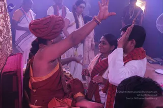 A photograph taken indoors under purple-tinted stage lighting. In the left foreground a seated figure wearing a red-orange turban, bare shoulder draped in orange-red cloth with gold trim, and a beaded rudraksha-style mala raises one arm high, palm open, toward two people on the right. On the right, a man in white kurta with a red-and-maroon shawl and a woman in cream and red sari with a red shawl also raise their palms in response. Behind them, several people in white and cream clothing stand, one woman wearing a pearl-like garland. A golden decorated panel is partly visible at the far left, and a fan and electronic equipment sit at the right edge. Photo credit overlay reads 2020 Sri Nithyananda Paramashivam.