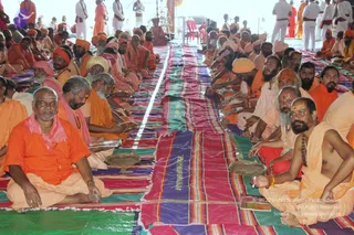 A large group of seated sadhus in saffron and orange robes sits in rows on colourful striped mats inside a tent. Many wear orange and yellow turbans or head cloths, and several have sacred ash marks on their foreheads. In the foreground, a man in orange with a pink shawl sits beside a bare-chested man in saffron cloth with a rudraksha mala and tilak, holding a small brass plate. Behind them, men in white clothing stand near the back of the tent and a red-cushioned chair is visible. A striped runner with the word a person is laid down the centre aisle.