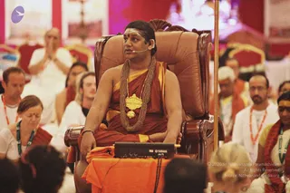 A seated man in an ornate brown throne-style chair at the centre, wearing a rust-red and saffron-orange draped garment, a heavy gold pendant on layered beaded necklaces, and a small microphone clipped near his face. A Yamaha mixer sits on a saffron cloth draped over his lap and armrest. Around him, seated devotees in white clothing and some wearing orange lanyards listen, one man with palms pressed together, and a woman in white with eyes closed. A red and pink backdrop with framed images and a tall gold pole fills the background. Appearance-based label: a person, an in_person candidate given the throne, saffron clothing, gold necklace and the page's note that he is present, with his own strong identifying cues being the ornate gold necklace and saffron garments.