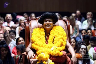 A seated man in a large brown leather armchair with carved wooden armrests sits centrally on a stage, wearing a rust-orange robe and a thick marigold garland of bright yellow flowers draped over his shoulders and chest, with a white-and-yellow flower tied at the lower garland. He has long dark hair swept upward, a red tilak on his forehead, a beaded rudraksha-style mala around his neck, and a small headset microphone near his face. He smiles warmly with his hands resting on the chair arms. Behind him a large audience of mostly adults fills the seating area, many smiling or watching attentively, some with cameras raised. A lit stage backdrop and purple logo appear in the upper corner.