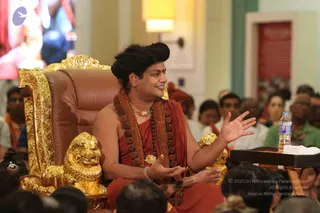 A seated man in saffron-orange robes sits on an ornate golden throne with a brown leather cushion and carved gold lion-head armrests, gesturing with both hands as he speaks. He wears a long rudraksha mala with a gold pendant necklace, and his long dark hair is swept up. Behind him a large screen shows colourful imagery. In the foreground the backs of audience members' heads are visible, along with a water bottle and white tissue on a wooden side table. Several seated devotees in orange and white clothing fill the background of a bright hall with cream walls and a red panel. Swamiji (probable): long swept-back hair, saffron robes, rudraksha mala and gold ornaments, seated on a throne during a discourse.