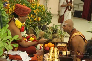 A photograph of a seated figure in a saffron-orange turban and saffron robe, wearing a thick garland of yellow and red flowers and rudraksha bracelets, smiling and gesturing toward a bald man with white forehead markings and rudraksha beads who sits facing him. In front of them is a wooden tray holding oranges, a red fruit, rudraksha mala strands, bangles, brass lamps and small packets. Potted green plants and rocks surround them, and a woman in a white sari with a rudraksha mala stands behind. The setting appears to be an indoor hall with a red curtain. The seated figure could be a person, based on the saffron turban, saffron robe, rudraksha bracelets and flower garland, though the appearance is not certain.