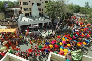Aerial view of a street procession seen from a building rooftop. Below, a crowd fills the road with many men in white and saffron-orange clothing carrying large colourful umbrellas in yellow, green, blue, pink and purple. Red flags are held aloft among the marchers. On the left, an ornate float with orange canopy and flower garlands carries a framed portrait. Behind, a white multi-tiered temple shrine with a tall spire rises above a white terrace where people stand. A white decorated vehicle with a digital display board and a banner moves through the procession. Trees line the street and more people watch from balconies above.