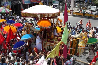 A decorated palanquin procession moves through a crowded street, carried by men in white dhotis. Mounted on the palanquin, a seated figure wears a red-brown turban and garland, with a white-clad attendant beside. Above them a white canopy with gold fringe and a row of hanging brass bells shelter the scene, and marigold and flower garlands drape the poles and frame. Many colourful umbrellas in orange, blue, red and green surround the palanquin. Red and green flags wave above the crowd, and uniformed police stand along the route. Onlookers in saffron, white and bright clothing press forward, many raising mobile phones to photograph, with a few saffron-robed sadhus visible at the upper right. Shops and a Gift Gallery storefront line the street behind them.